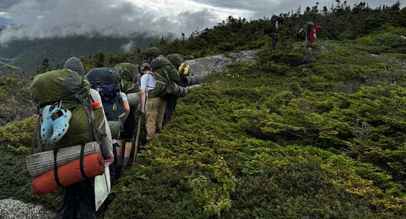 A group of students wearing backpacks hike along a path in a open green area. 